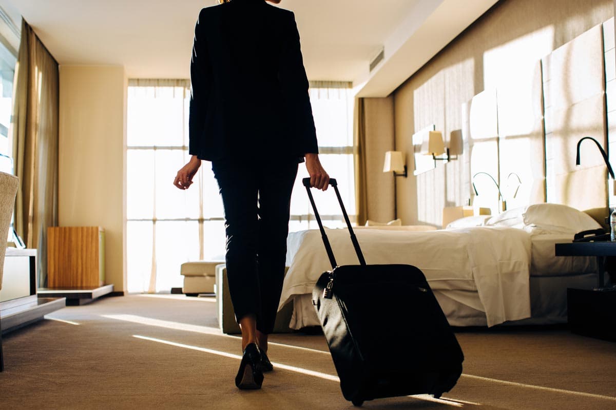 A woman wearing black is shown from behind as she walks into a hotel room pulling a suitcase behind her.