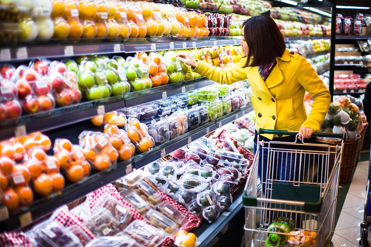 A woman wearing a yellow jacket browses produce in a grocery store.